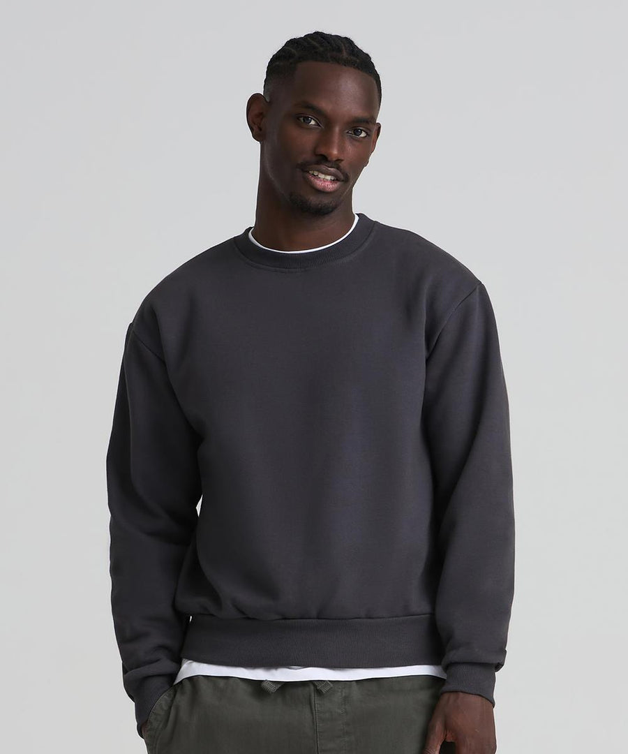 A man faces the camera wearing the AWDis Just Hoods Vision heavyweight sweatshirt in dark grey over a white shirt and green trousers, set against a plain, light background.