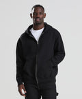 A man faces the camera with his right hand in his pocket, wearing the AWDis Just Hoods Vision heavyweight zoodie over a white T-shirt and black trousers against a plain light grey background.