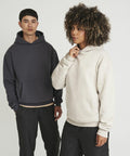 Two individuals stand side-by-side wearing AWDis Just Hoods JH120 hoodies; one in a dark gray hoodie, the other in a light beige hoodie. Both have their hands placed on or near their hoodies' pockets. Background is plain white.