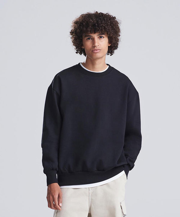 A person with curly hair stands against a plain background, wearing the AWDis Just Hoods Signature heavyweight sweatshirt in black over a white T-shirt and light-coloured trousers, with one hand in their pocket.
