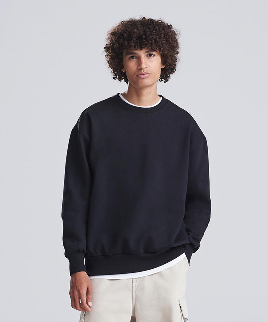 A person with curly hair stands against a plain background, wearing the AWDis Just Hoods Signature heavyweight sweatshirt in black over a white T-shirt and light-coloured trousers, with one hand in their pocket.