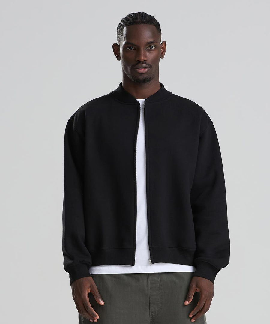 A man stands facing forwards against a plain background, wearing the AWDis Just Hoods Signature heavyweight bomber sweatshirt in black over a white shirt and olive green trousers.