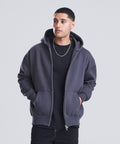 A person wearing the AWDis Just Hoods Signature heavyweight zoodie in dark grey over a black t-shirt stands with hands in the pockets, looking slightly to the side against a plain light grey background.