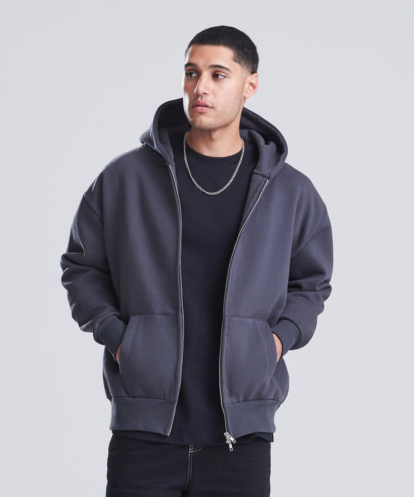 A person wearing the AWDis Just Hoods Signature heavyweight zoodie in dark grey over a black t-shirt stands with hands in the pockets, looking slightly to the side against a plain light grey background.