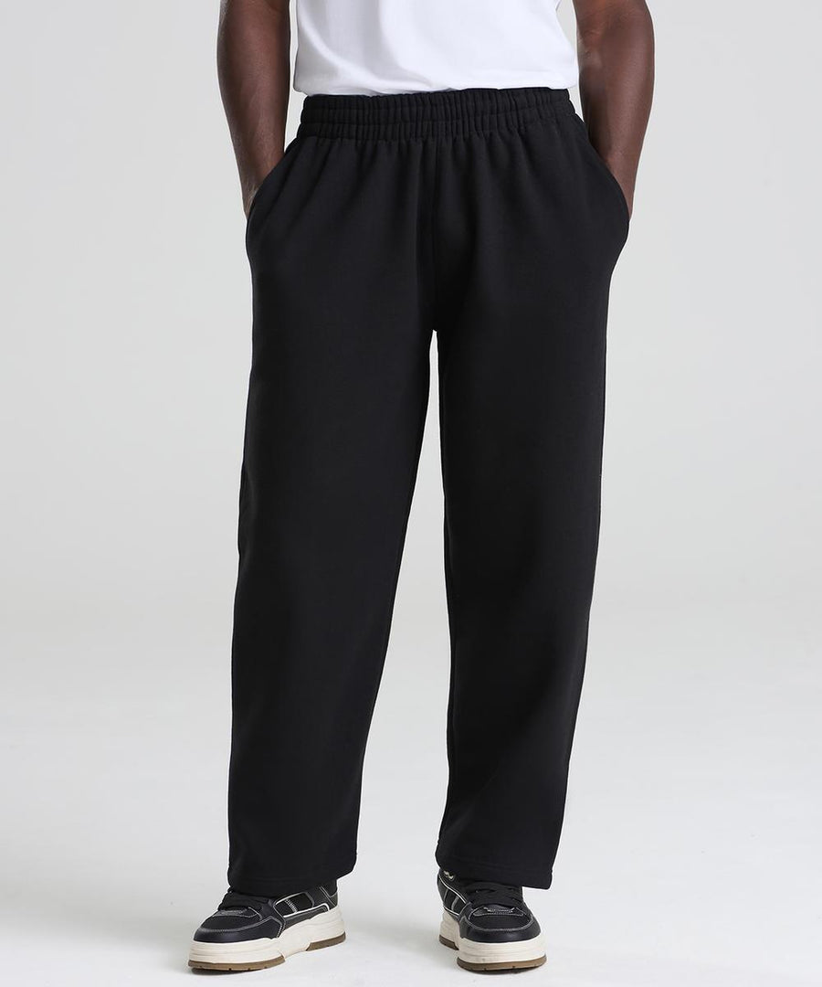 Only the lower half of a person is visible, wearing AWDis Just Hoods Signature heavyweight joggers in black, paired with black and white trainers and a white shirt, standing on a white background with hands in pockets.