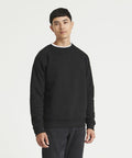 A man stands against a white background, wearing a black long-sleeve JH130 sweatshirt from AWDis Just Hoods and dark pants.