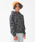 A child with curly hair wears an orange JH14J by AWDis Just Hoods, a camo-pattern hoodie, and light gray sweatpants, standing against a plain white background.
