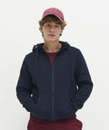 A person with curly hair wears a red cap, a navy blue AWDis Just Hoods JH150 hoodie with a zipper, and maroon pants. They are looking directly at the camera against a plain white background.