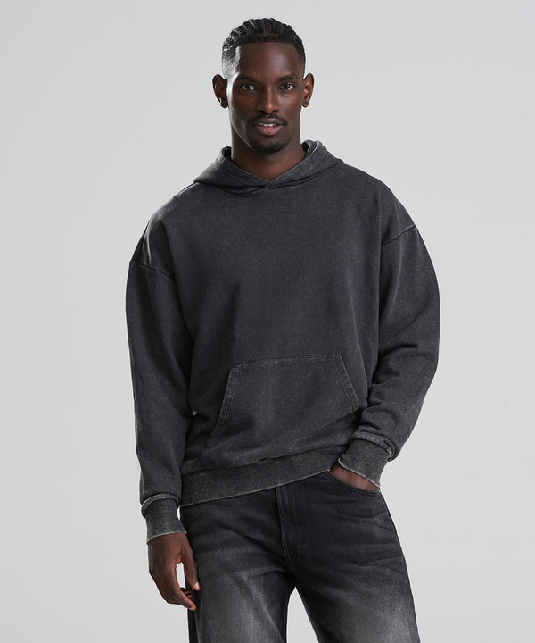A man with short cornrows stands against a plain light background, wearing the AWDis Just Hoods Vintage washed hoodie in dark grey and black jeans, with one hand in his pocket and the other at his side.