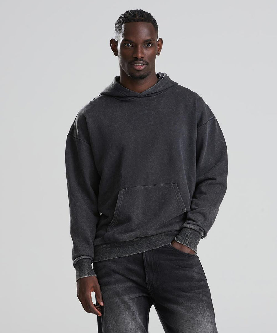 A man with short cornrows stands against a plain light background, wearing the AWDis Just Hoods Vintage washed hoodie in dark grey and black jeans, with one hand in his pocket and the other at his side.