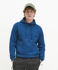 A person with curly hair wearing an AWDis Just Hoods JH201, black pants, and a green cap, standing against a plain white background.
