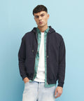 A person stands against a blue background wearing a black JH250 hoodie by AWDis Just Hoods over a green shirt and a white t-shirt, with light blue jeans.
