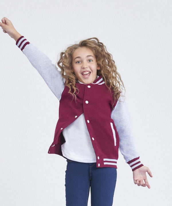 A smiling child with curly hair, wearing an AWDis Just Hoods JH43J varsity jacket, white shirt, and blue pants, raises one arm enthusiastically in the air.