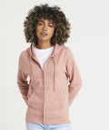 Person with curly hair wearing an AWDis Just Hoods JH50F pink zip-up hoodie over a white shirt, with hands in the front pockets, standing against a plain white background.