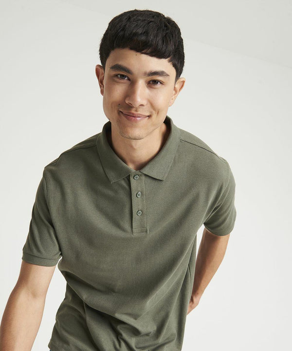 A person with short, dark hair is standing and smiling, wearing a green JP100 polo shirt from AWDis Just Polo's.