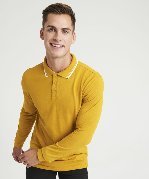 A person with short hair and a smile is wearing a mustard yellow AWDis Just Polo's JP103 long-sleeve polo shirt, standing against a plain white background.