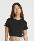 A woman with long hair and tattoos on her arms wears a black JT006 T-shirt by AWDis Just T's and light blue jeans, standing against a plain white background.