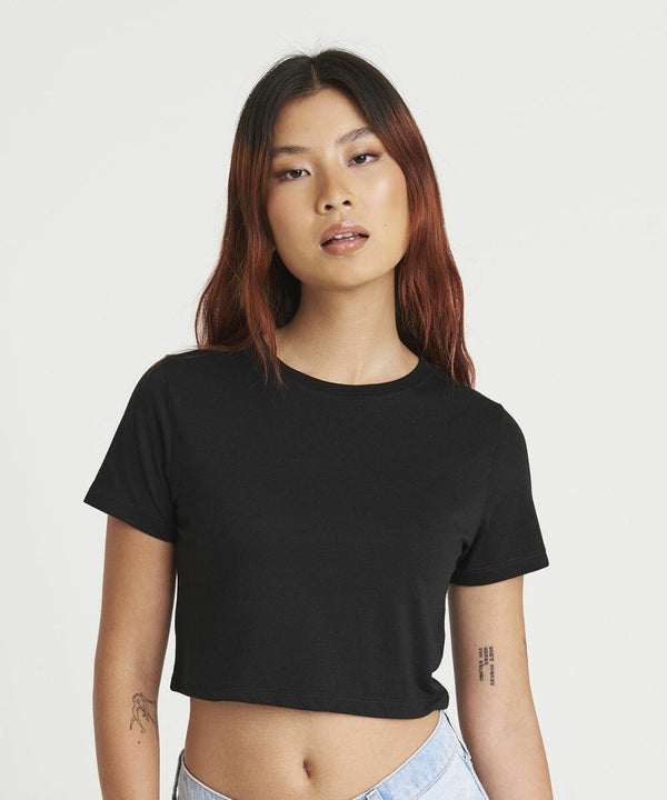 A woman with long hair and tattoos on her arms wears a black JT006 T-shirt by AWDis Just T's and light blue jeans, standing against a plain white background.