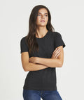 A person with long dark hair, wearing an AWDis Just T's JT01F t-shirt and blue jeans, stands with arms crossed against a white background.