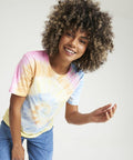 A person with curly hair, wearing a JT022 T-shirt by AWDis Just T's and jeans, is smiling and leaning slightly forward.