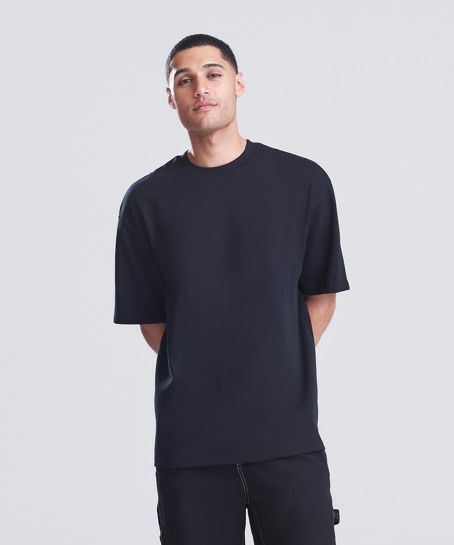 A person stands facing forwards with their hands behind their back, wearing the AWDis Just T's Signature heavyweight T-shirt and black trousers against a plain light grey background.