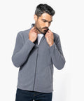 A man with short, dark hair adjusts the collar of his light gray Kariban K9102 zip-up jacket. He is wearing a white t-shirt underneath and dark jeans. The background is plain white.