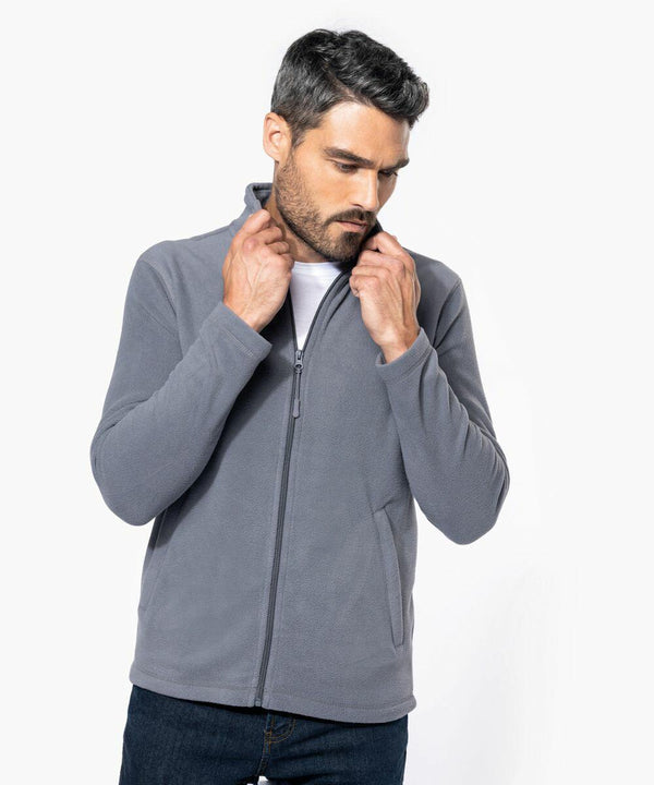 A man with short, dark hair adjusts the collar of his light gray Kariban K9102 zip-up jacket. He is wearing a white t-shirt underneath and dark jeans. The background is plain white.