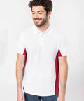 A man with a beard and short hair wearing a white Kariban polo shirt with red side panels, model KB232, stands against a plain background, looking down and smiling.