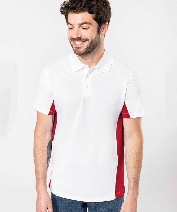 A man with a beard and short hair wearing a white Kariban polo shirt with red side panels, model KB232, stands against a plain background, looking down and smiling.
