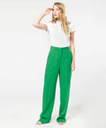 A person stands against a white background wearing a white KB251 by Kariban and high-waisted green trousers, with one hand raised to their forehead.