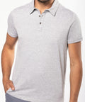 A person wearing a Kariban KB262 light gray short-sleeve polo shirt with a three-button placket. The person is standing with one hand in their pocket.