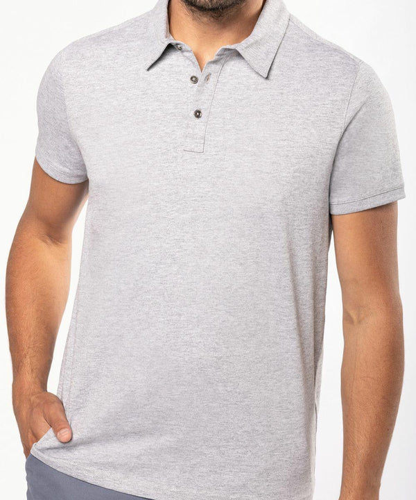 A person wearing a Kariban KB262 light gray short-sleeve polo shirt with a three-button placket. The person is standing with one hand in their pocket.