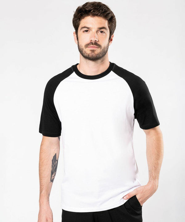A man with short dark hair and a beard is wearing a white Kariban KB330 T-shirt with black sleeves. He has a tattoo on his left forearm and stands with one hand in his pocket against a plain background.