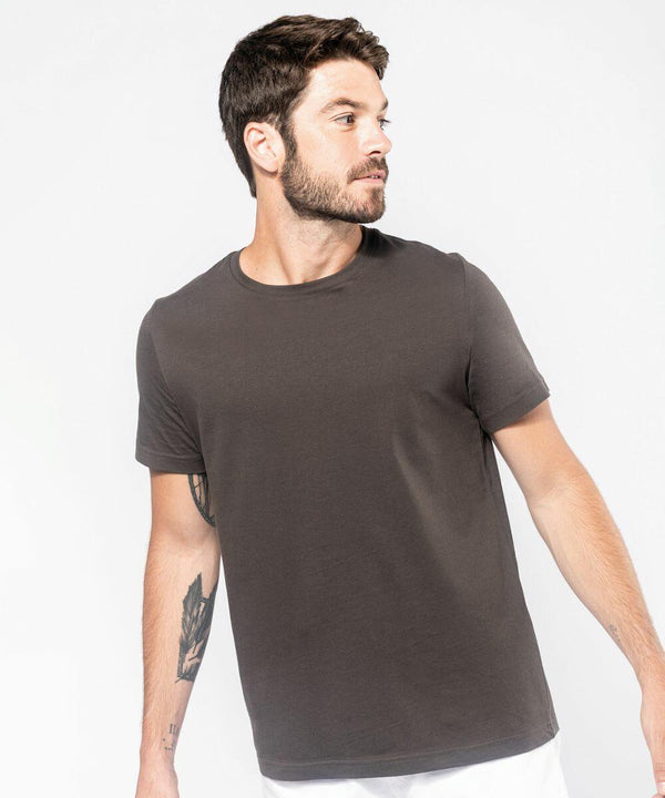 A man with short brown hair, facial hair, and a tattoo on his arm is wearing a Kariban KB356 t-shirt and looking to the side against a light background.
