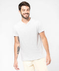 A man in a light gray Kariban KB357 V-neck T-shirt and light-colored pants is smiling and looking to his left. He has short dark hair and a beard. His right arm has visible tattoos.