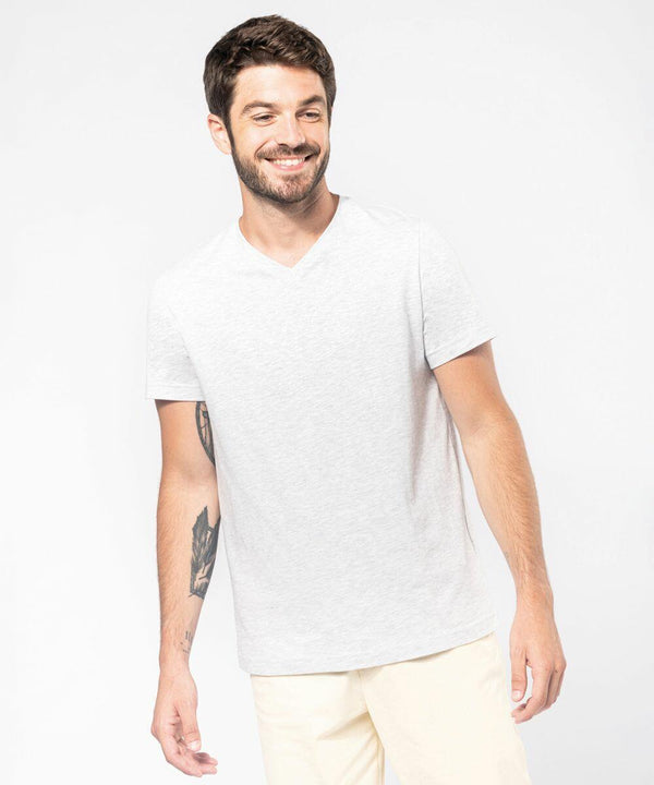 A man in a light gray Kariban KB357 V-neck T-shirt and light-colored pants is smiling and looking to his left. He has short dark hair and a beard. His right arm has visible tattoos.