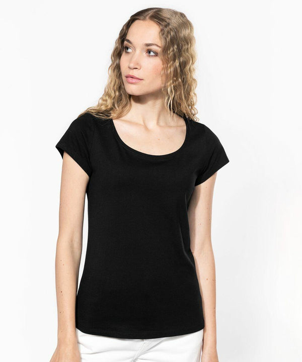 A woman with long wavy hair is wearing a plain black short-sleeved Kariban KB384 and white bottoms, standing against a white background. She is facing slightly to the side, looking off into the distance.