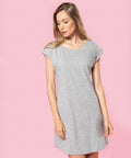 A person with long hair wears a short-sleeved, knee-length gray dress (KB388 by Kariban) and stands against a pink background.