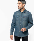 Man with dark hair and beard wearing a Kariban KB519 button-down denim shirt and black pants, looking to his left, against a white background.