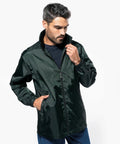 A man with short hair and a beard adjusts the zipper of his dark green Kariban KB616 windbreaker while looking down. He is wearing a black shirt and blue jeans.