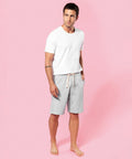 A man stands barefoot on a pink background, wearing a white Kariban T-shirt and gray KB710 drawstring shorts, with hands in his pockets.