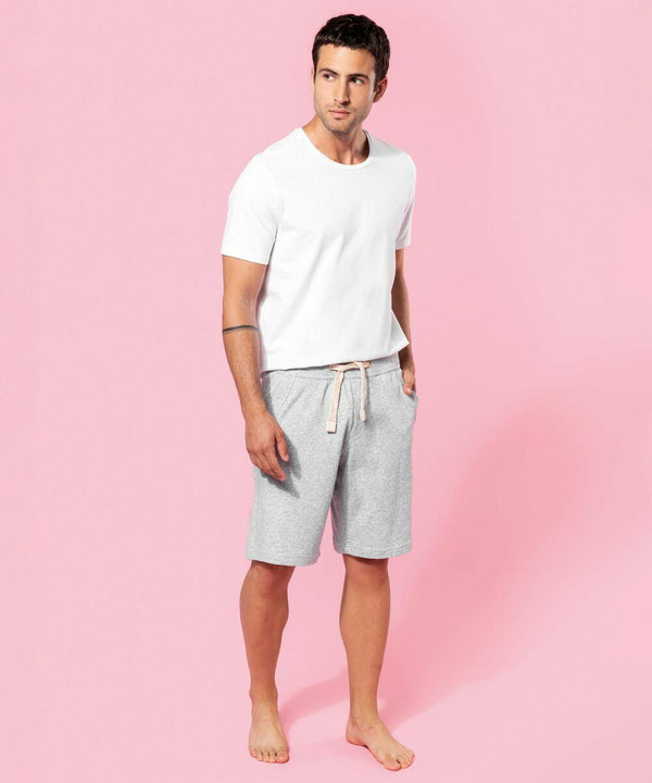 A man stands barefoot on a pink background, wearing a white Kariban T-shirt and gray KB710 drawstring shorts, with hands in his pockets.