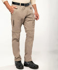 A person wearing Kariban KB785 beige cargo pants with zip-off legs stands with hands in pockets, paired with a light-colored shirt and black hiking shoes.