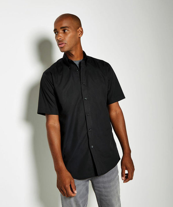 A person standing against a light gray background, wearing a short-sleeve black button-up shirt by Kustom Kit and gray pants, looking to the side. The shirt is the KK100 model.