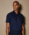 A person wearing a dark blue, short-sleeved KK102 shirt from Kustom Kit stands against a plain, beige background.