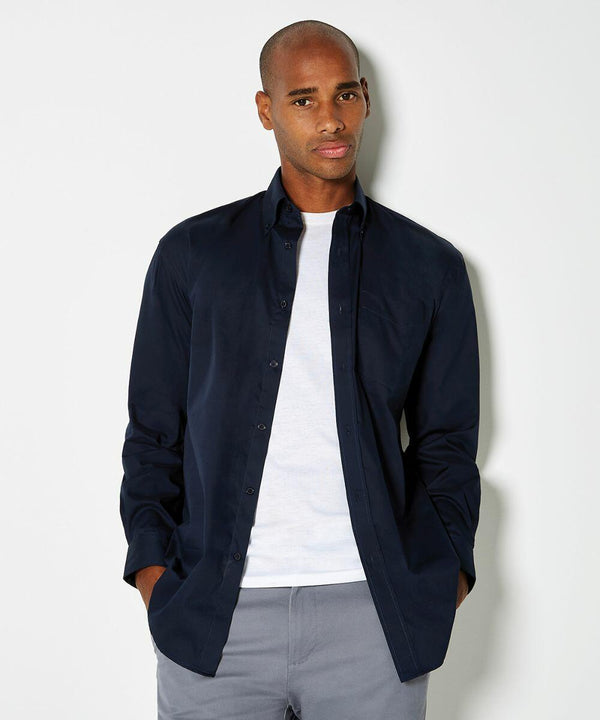 A man wearing a dark navy Kustom Kit KK105 button-down shirt over a white t-shirt and gray pants, standing against a plain light-colored background.