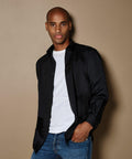 A man stands against a beige background wearing a Kustom Kit KK140 over a white T-shirt and blue jeans, looking directly at the camera.