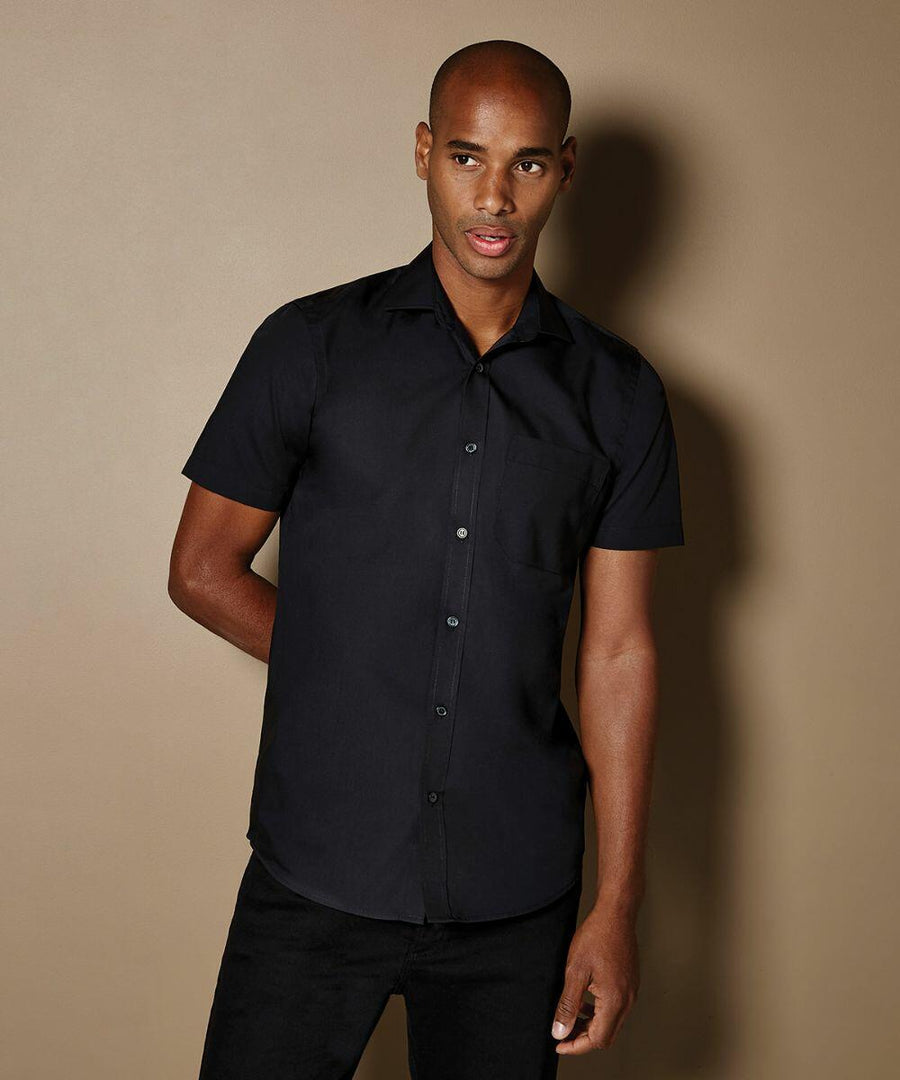 A man wearing a black short-sleeve button-up KK141 shirt by Kustom Kit stands against a plain brown background, looking slightly to the right.