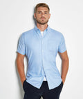 A man with short hair and a beard wears a light blue Kustom Kit KK183 short-sleeve button-up shirt and dark pants. He stands with his hands in his pockets against a white background.