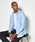 A man wearing a light blue KK184 dress shirt by Kustom Kit over a white t-shirt and dark pants is seated on a stool, looking to his left against a plain white background.